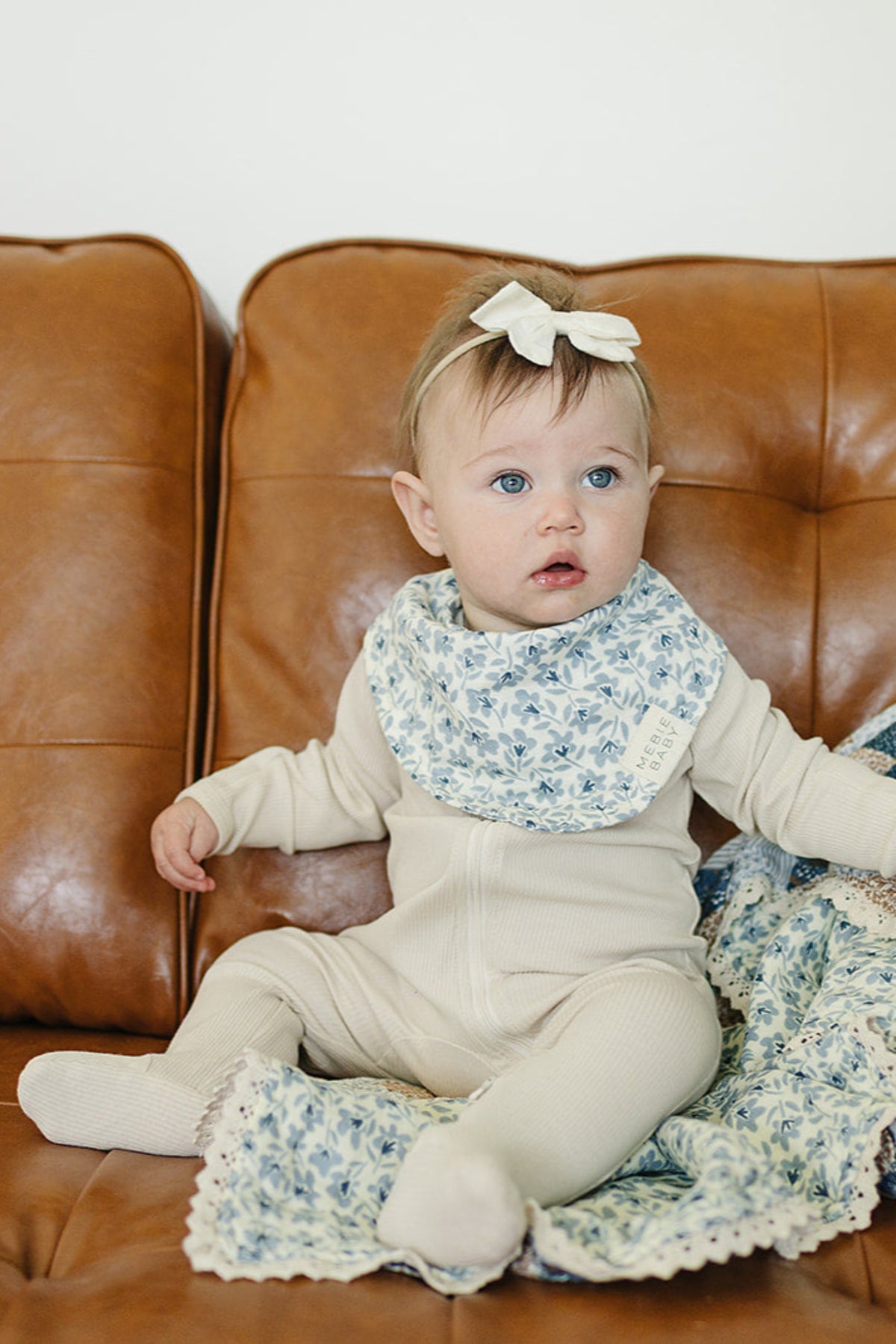 A baby wearing a blue floral bib and cream outfit sits on a brown couch, with a matching blanket.