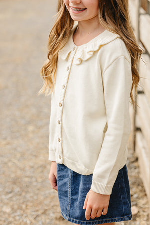 A young girl wearing a cream ruffle knit cardigan with wooden buttons stands outdoors, showcasing her casual outfit paired with a denim skirt.
