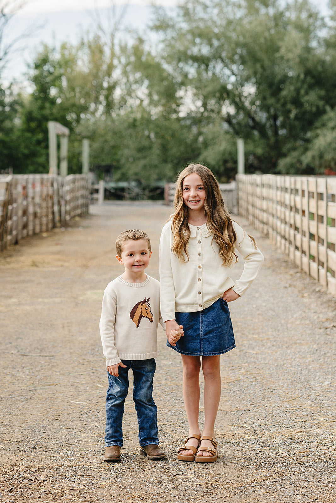 A young girl in a cream cardigan and denim skirt stands with a boy in a horse-embroidered sweater and jeans, both smiling on a rustic path surrounded by greenery.