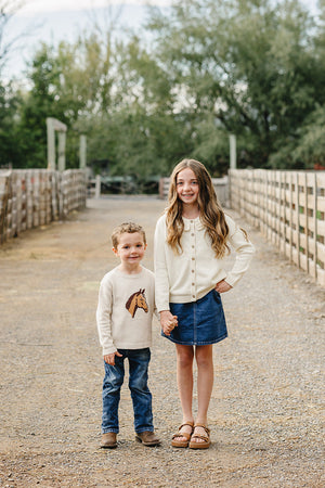 A young girl in a cream cardigan and denim skirt stands with a boy in a horse-embroidered sweater and jeans, both smiling on a rustic path surrounded by greenery.