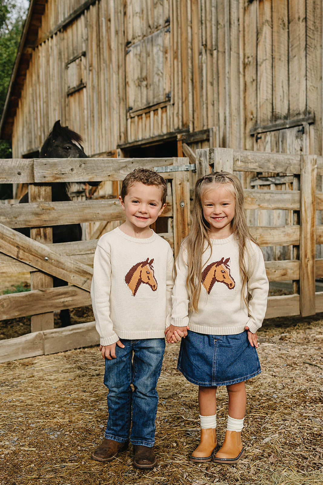 Two children stand outside a wooden barn, wearing cozy cream sweaters featuring a horse design.
