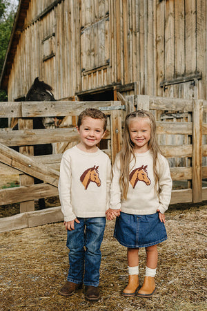 Two children stand outside a wooden barn, wearing cozy cream sweaters featuring a horse design.