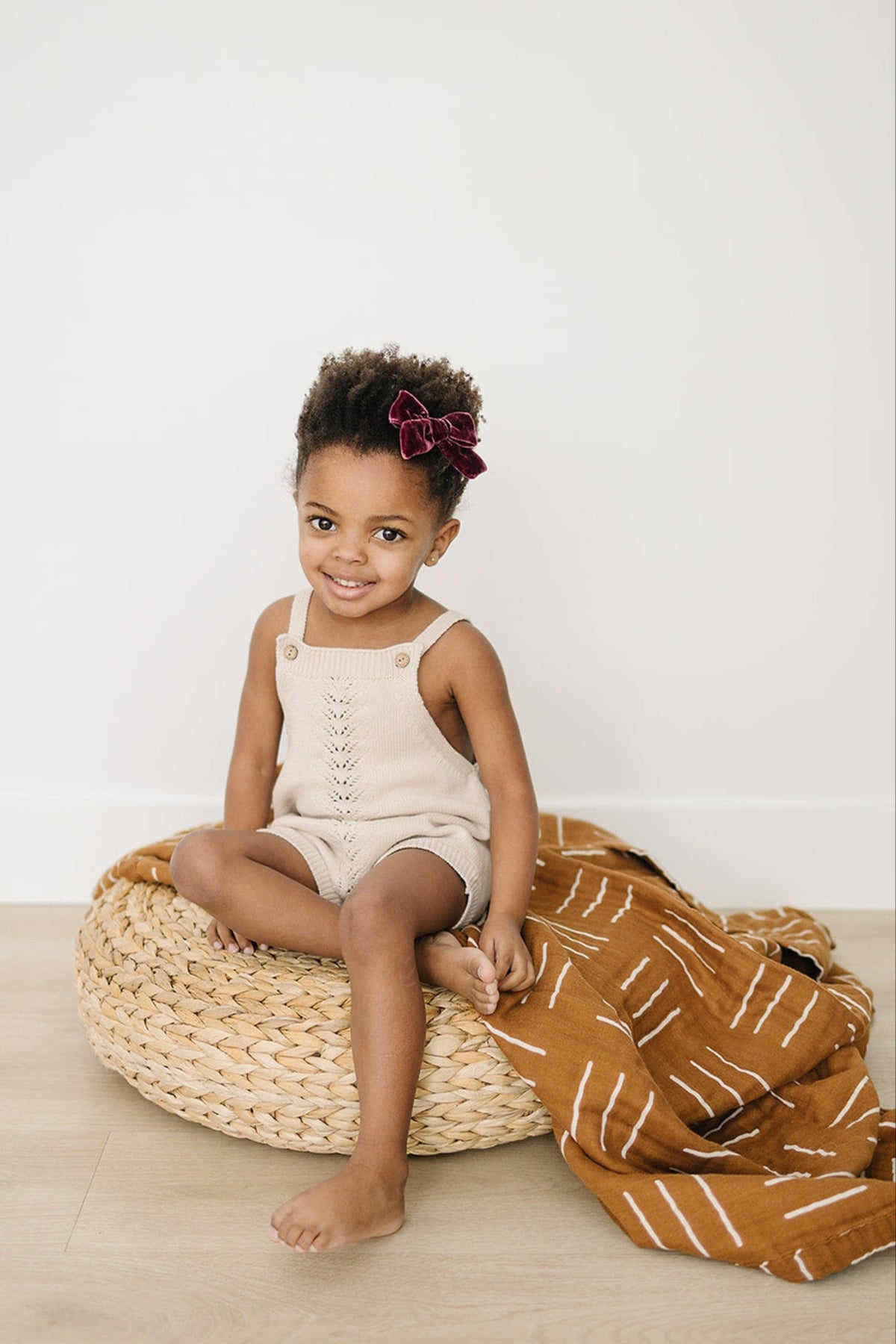 A child wearing a beige romper with a burgundy bow sits on a woven cushion beside a mustard quilt.