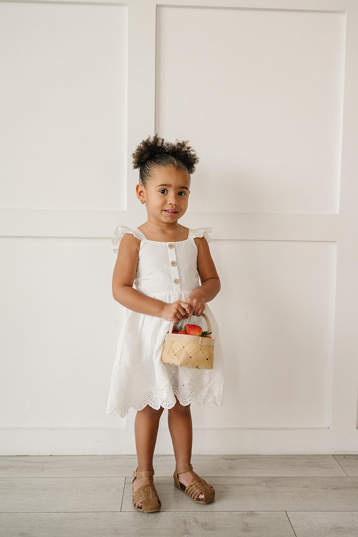 A young girl stands against a white wall, wearing a white eyelet cotton dress with ruffled straps and a scalloped hem.