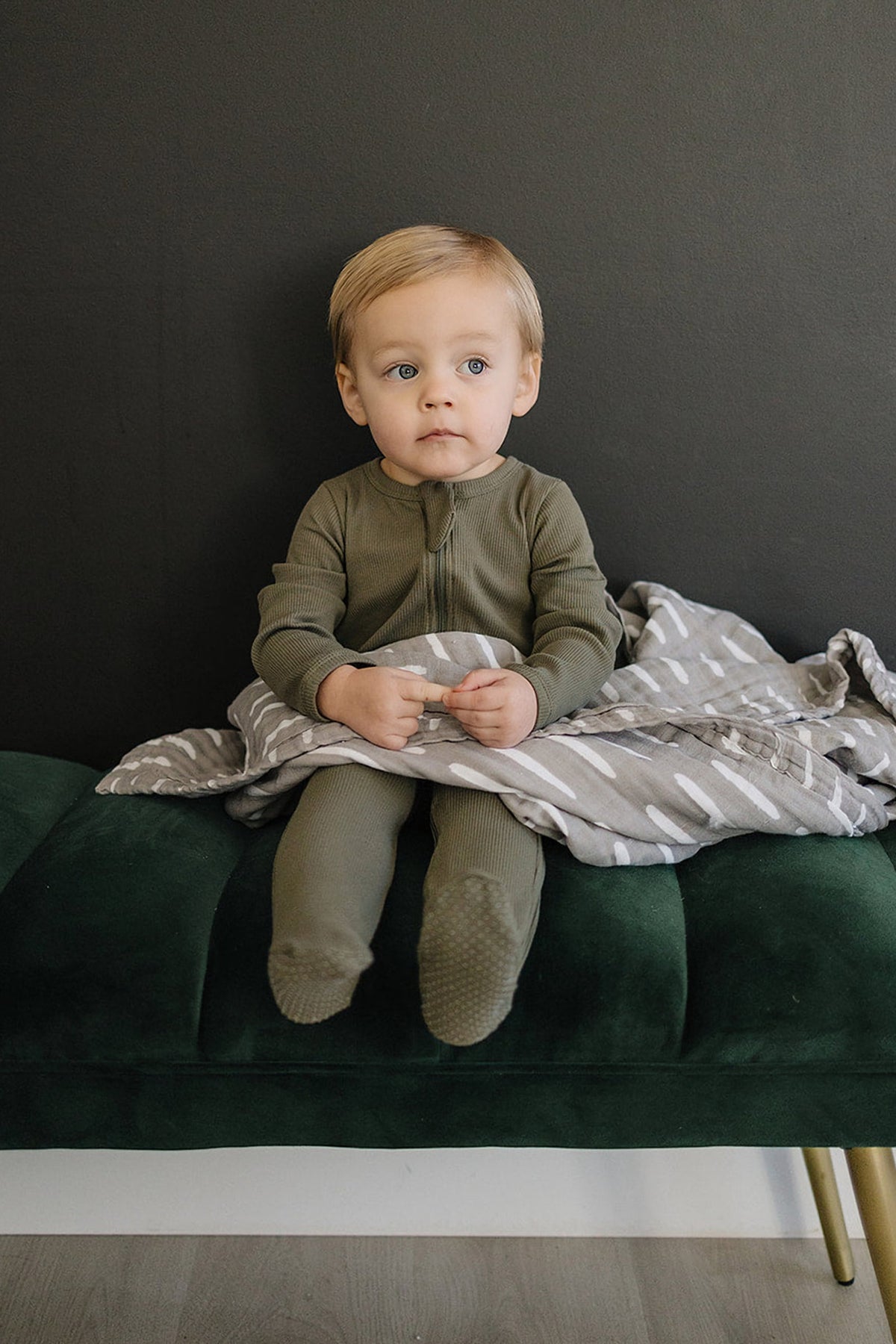 A toddler in green ribbed footed pajamas sits on a dark green bench with a patterned gray blanket.