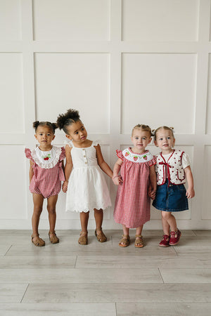 Four young children stand together, showcasing colorful outfits. Two girls wear gingham rompers with strawberry accents, while another sports a white dress, and a girl in a denim skirt completes.