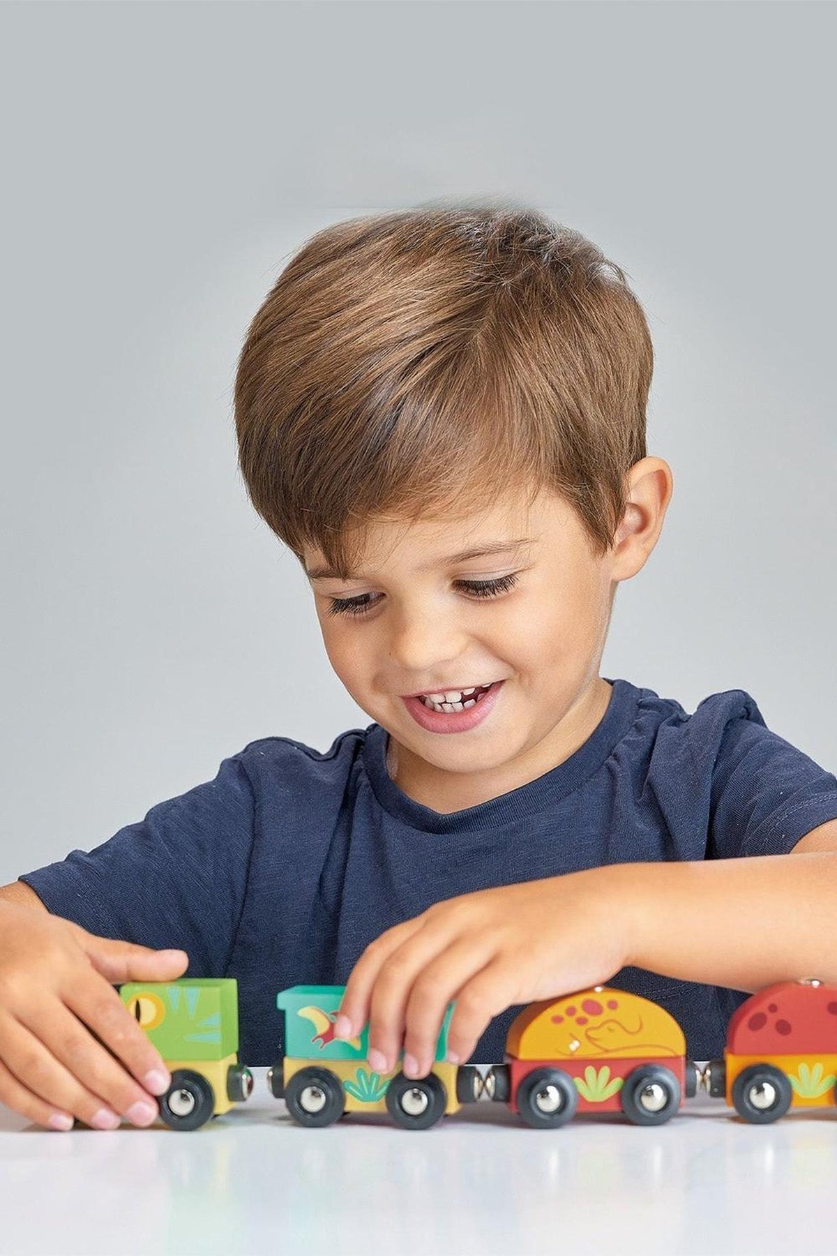 A young boy with short brown hair smiles while playing with a colorful wooden train set. The train features vibrant carriages, including a dinosaur-themed design, promoting imaginative play.