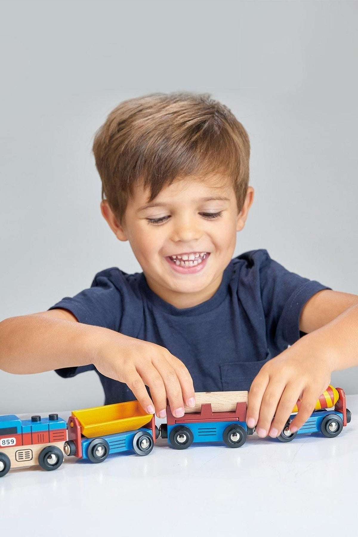 A young boy smiles as he plays with colorful wooden toy trains, featuring different carriages and vibrant designs, showcasing a fun, engaging play experience with sustainable materials.