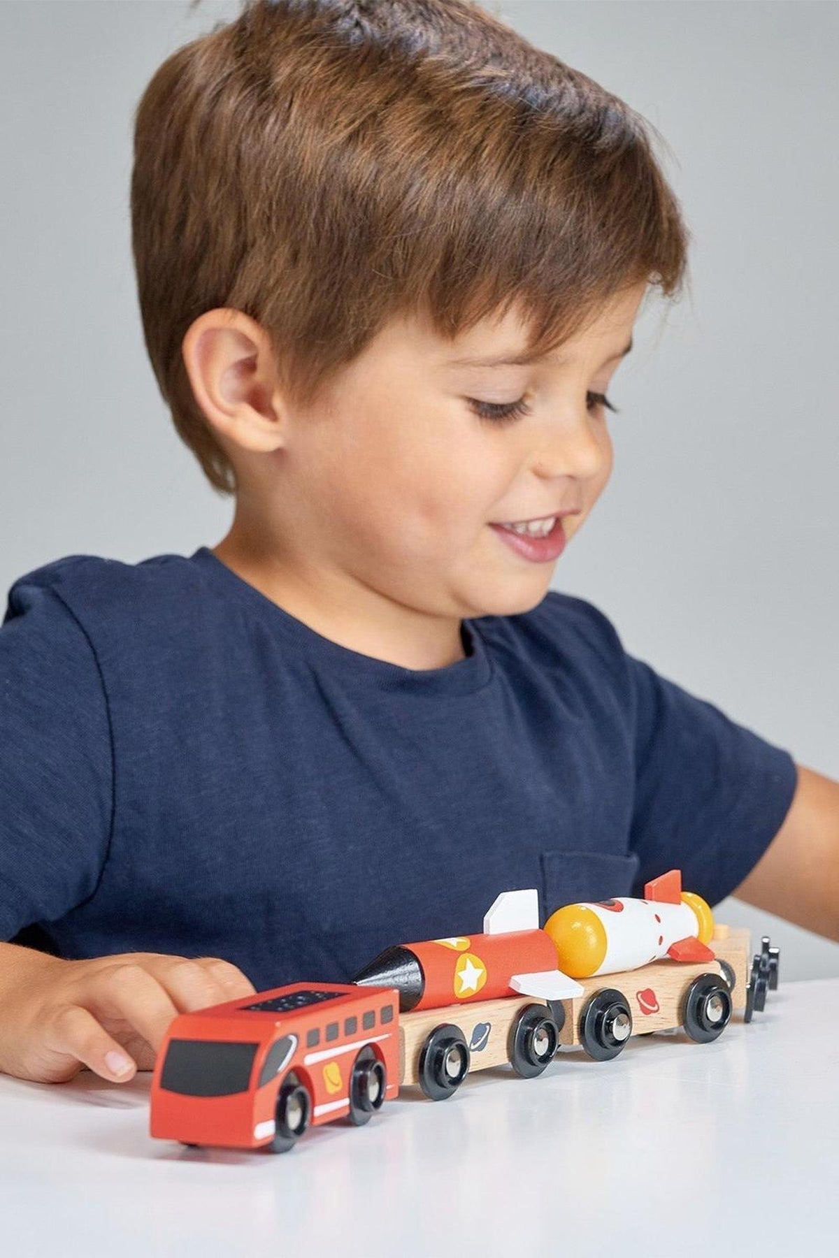 A young boy smiles while playing with a colorful wooden Space Rocket Train, featuring bright red and orange elements. The train includes multiple carriages and a detailed engine design.