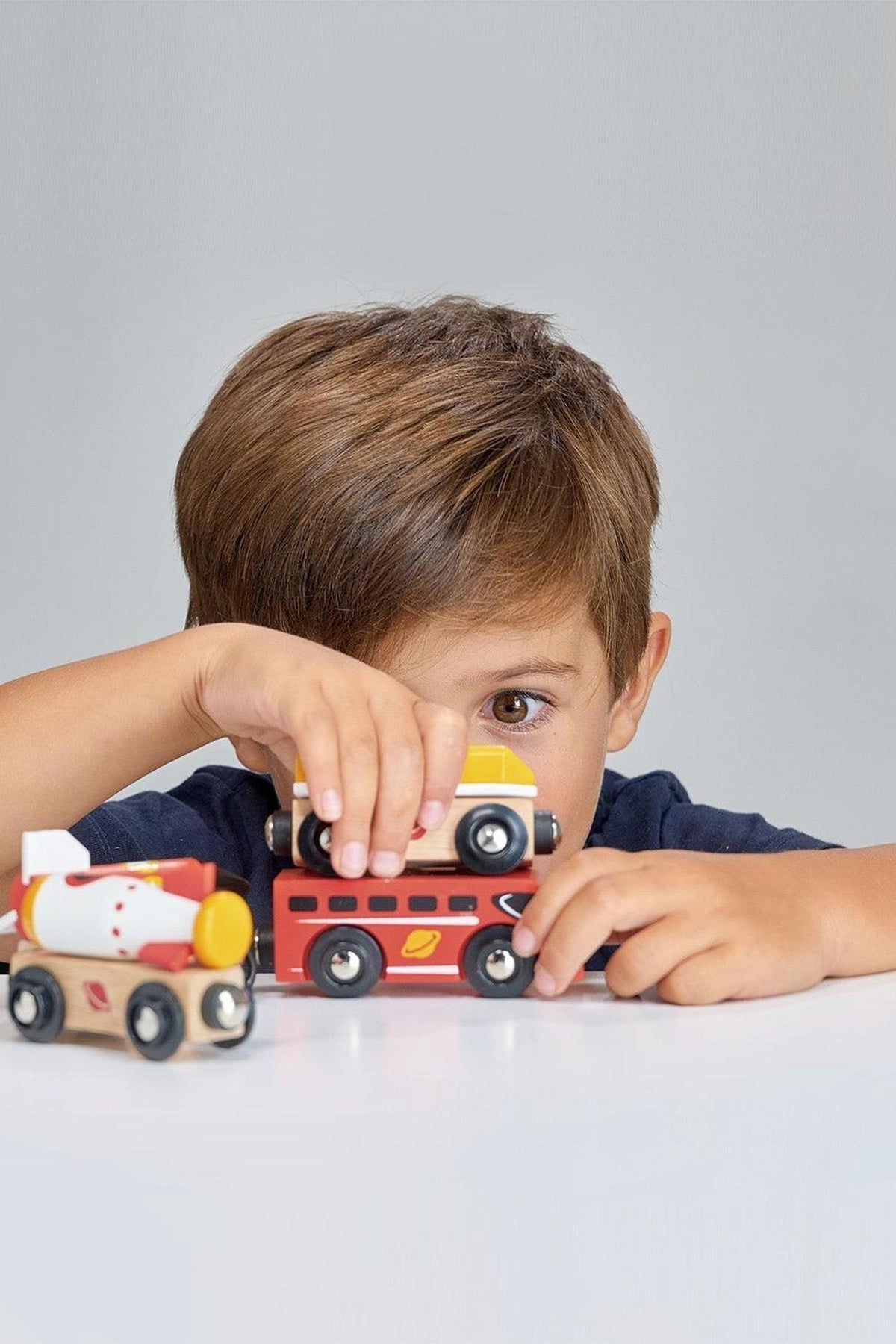 A young boy plays with colorful wooden toy trains, including a rocket train and a cargo train, showcasing bright red, yellow, and white colors against a neutral background.