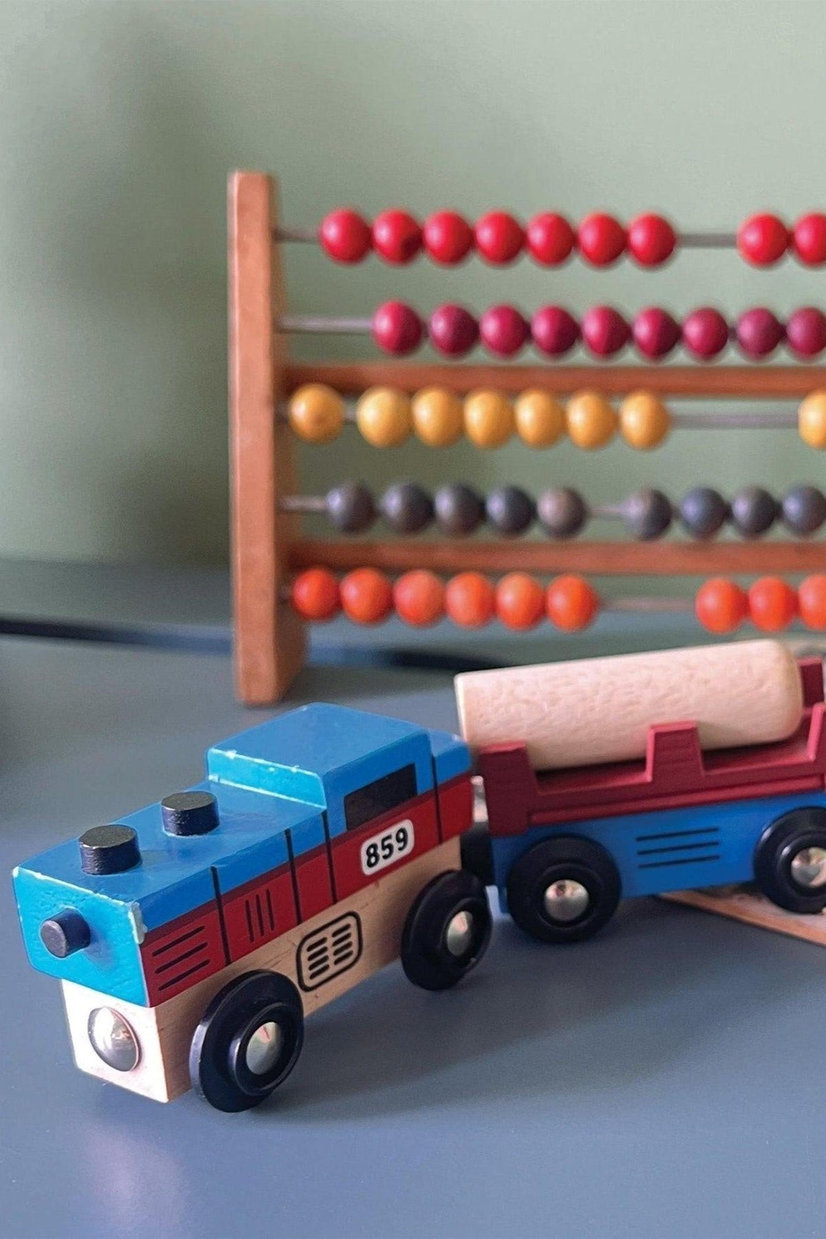 A colorful wooden toy train set featuring a blue engine and a red cargo car with a wooden log. The background includes a vibrant abacus with multicolored beads.