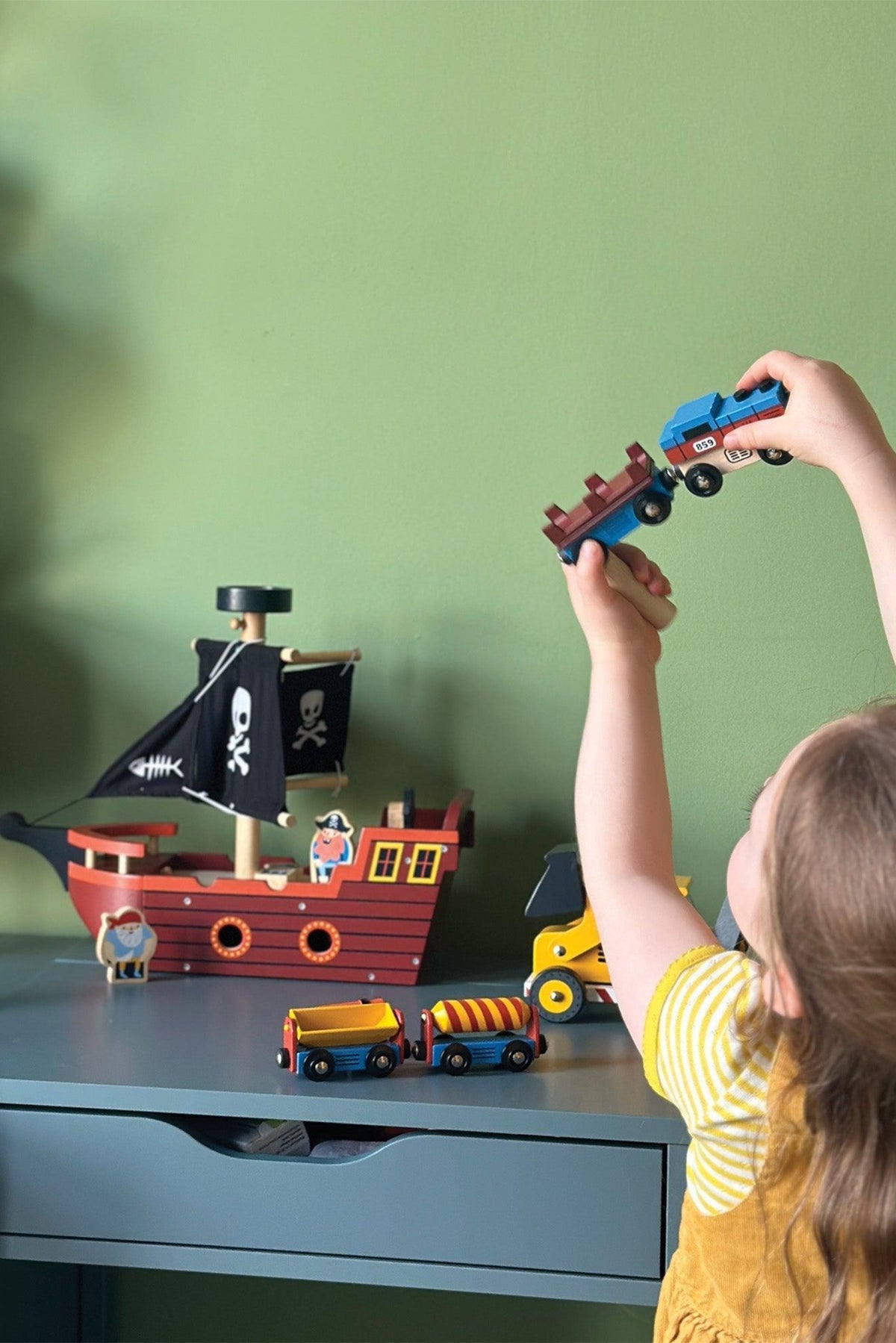 A child holds a wooden toy truck while playing near a pirate ship toy with a black flag, colorful train cars, and figurines on a green wall backdrop.