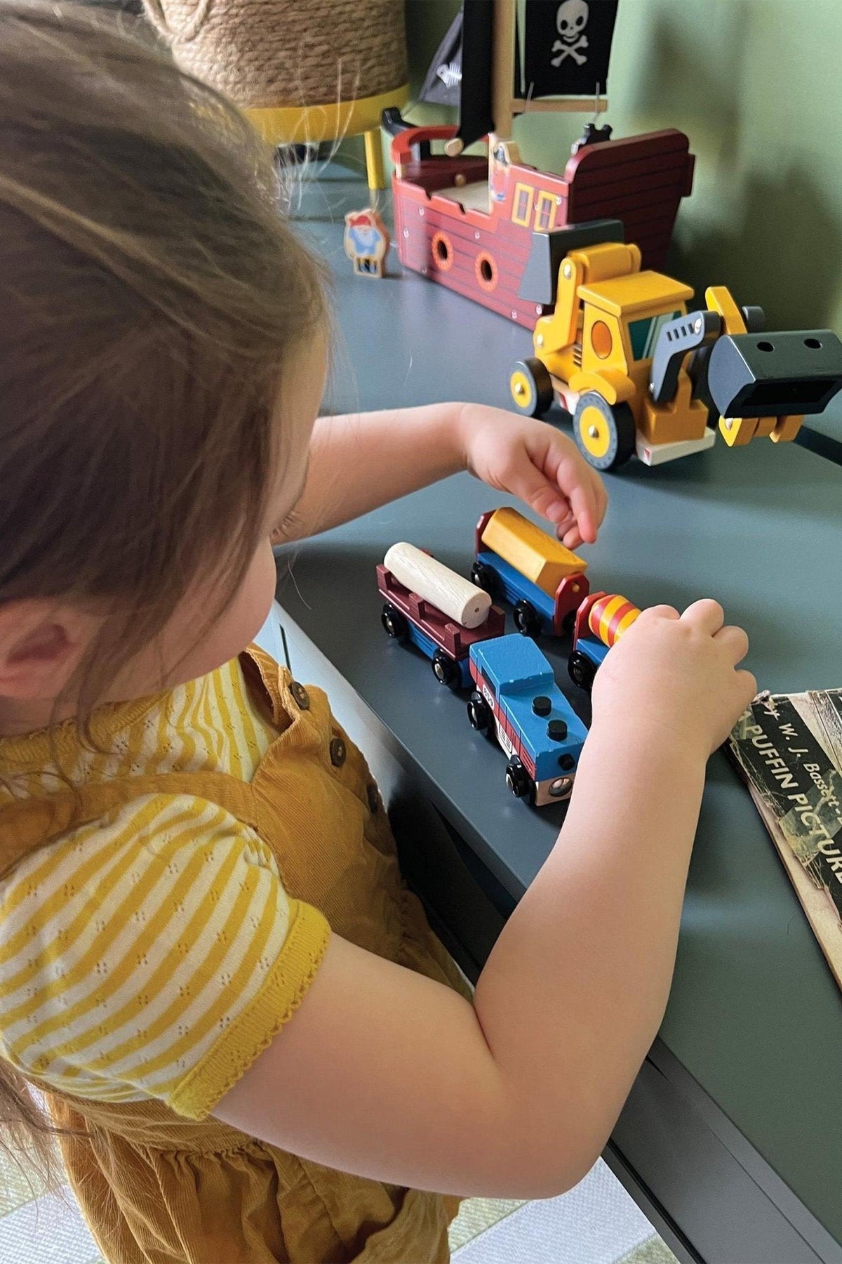 A child plays with colorful wooden toy trains on a table, featuring different carriages and vibrant designs. The setting includes playful elements like a pirate ship and construction vehicle.