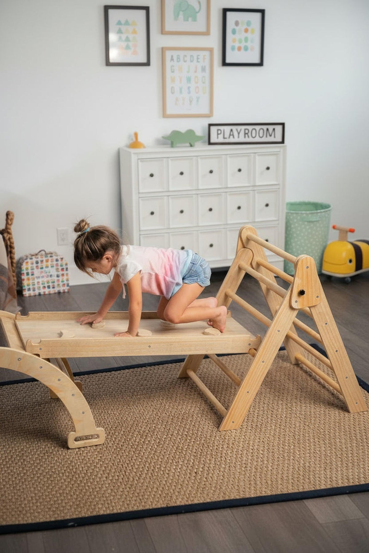 A child climbs on a wooden Pikler triangle playset in a bright playroom, featuring a soft area rug and colorful wall art.