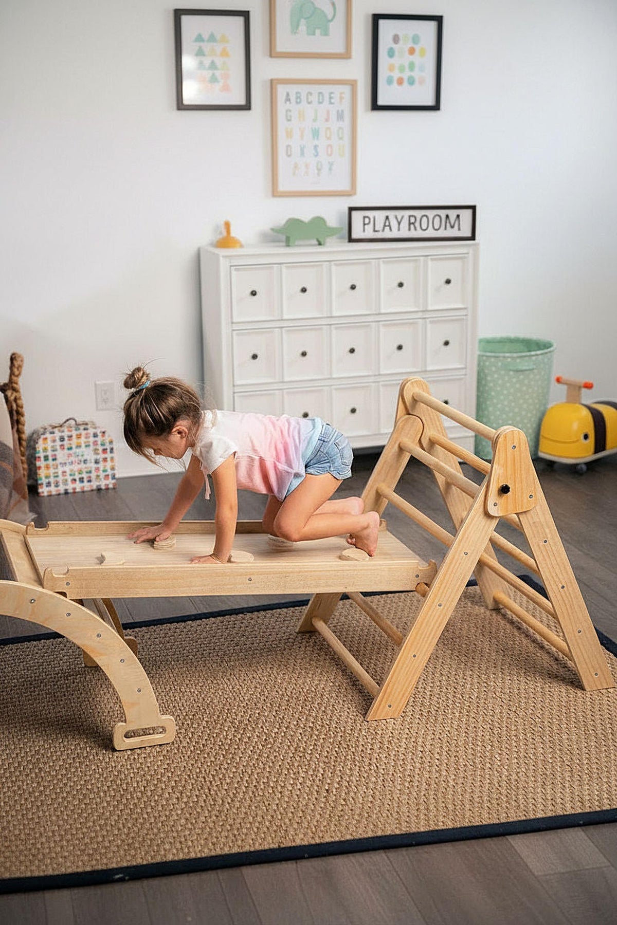 A child climbs on a wooden Pikler triangle playset in a bright playroom. The modular structure features a natural finish, promoting active play and exploration in a safe environment.