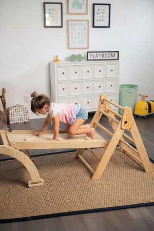 A child climbs on a wooden Pikler triangle playset in a bright playroom. The modular structure features a natural finish, promoting active play and exploration in a safe environment.