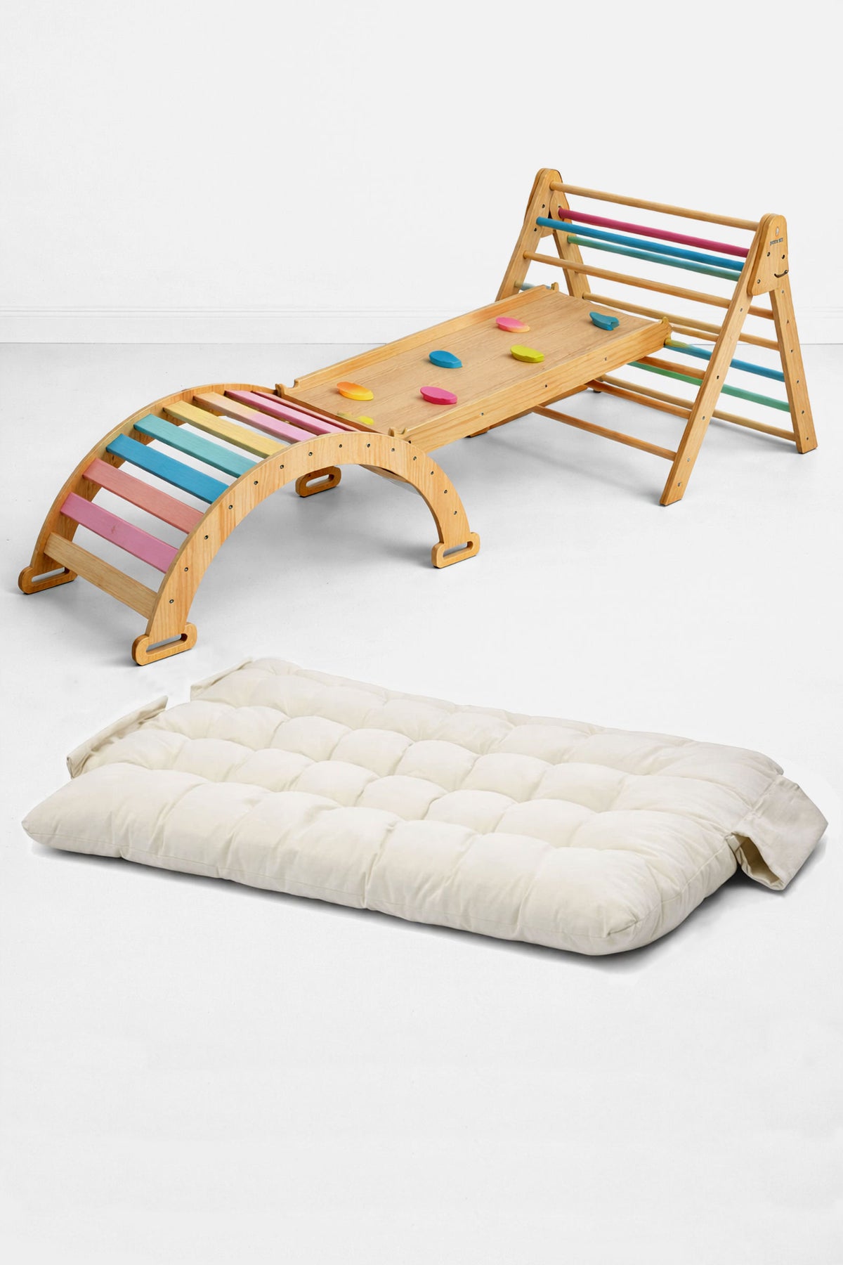 A wooden Pikler triangle playset features colorful climbing rungs and a slide, accompanied by a thick, cream-colored cushion. This sustainable, eco-friendly setup is ideal for active toddlers.