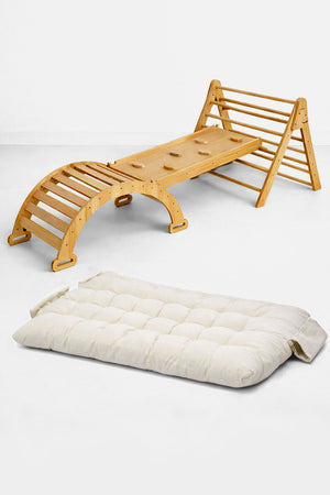 A wooden Pikler triangle playset with a slide and rocker features a natural finish, paired with a thick, cream-colored cushion. Ideal for indoor active play and comfort for toddlers.
