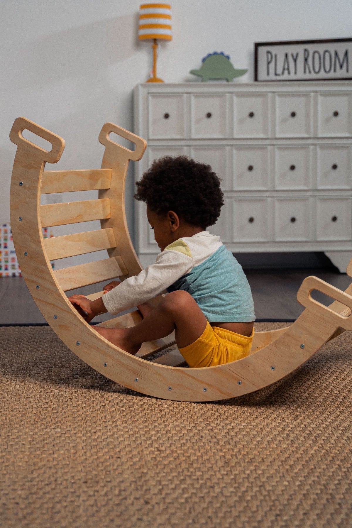 A child sits on a wooden rocker, wearing a colorful outfit, within a cozy playroom setting. The rocker features a smooth, curved design and is made from sustainable materials.