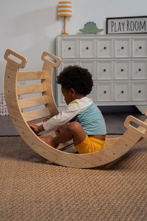A child sits on a wooden rocker, wearing a colorful outfit, within a cozy playroom setting. The rocker features a smooth, curved design and is made from sustainable materials.