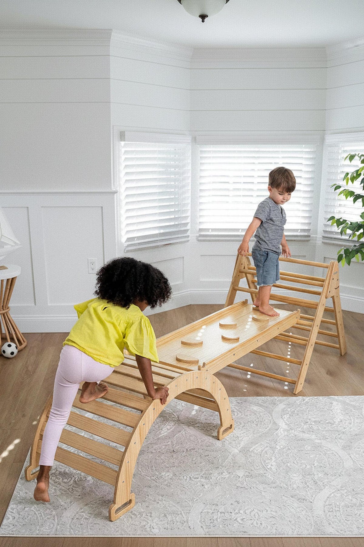 Two children play on a wooden Pikler triangle playset in a bright, airy room. One child climbs up, while the other balances on a bridge, showcasing the sets natural wood.