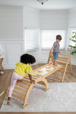 Two children play on a wooden Pikler triangle playset in a bright, airy room. One child climbs up, while the other balances on a bridge, showcasing the sets natural wood.