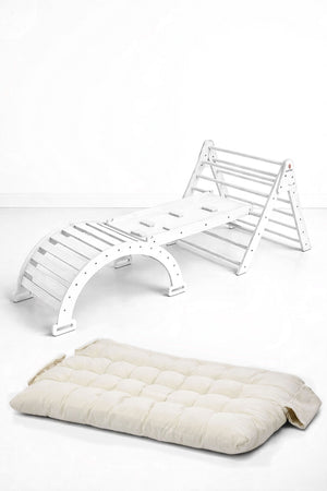 The image features a white wooden Pikler triangle playset alongside a thick, cream-colored cushion. This eco-friendly, modular design promotes active play and comfort for toddlers and young children.