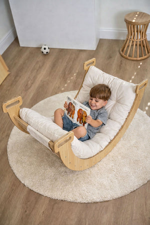 A young child sits comfortably in a wooden rocker, reading a book. The rocker features a soft, cushioned seat and is positioned on a plush.