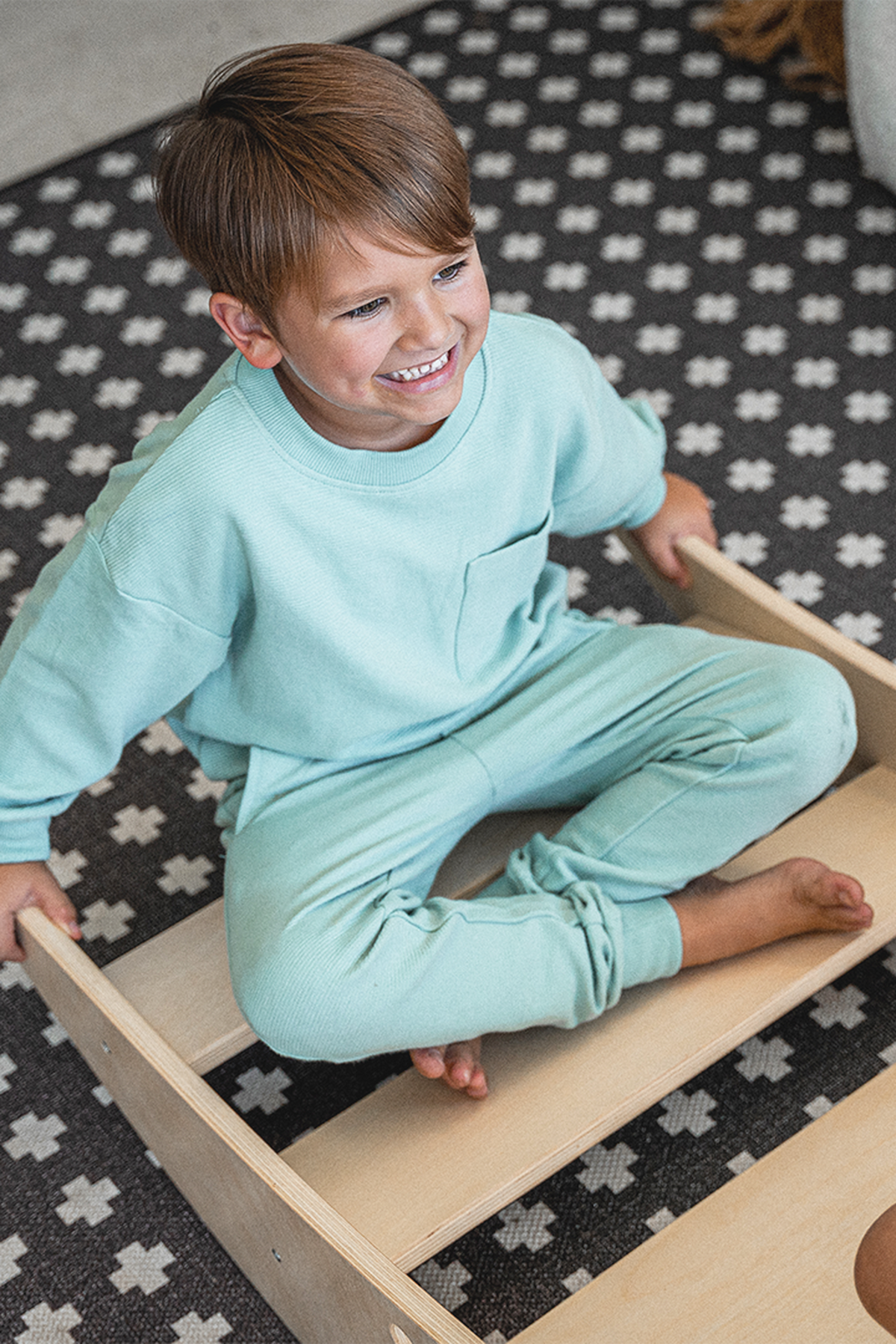 Two children play together on a light-colored wooden climbing bridge, smiling and sitting cross-legged. The setting features a patterned rug, enhancing a cozy and playful atmosphere.