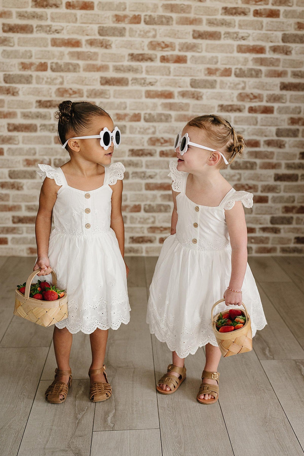 Two young girls wearing white eyelet cotton dresses stand side by side, each holding a basket of strawberries. They sport oversized sunglasses and are set against a brick wall backdrop.