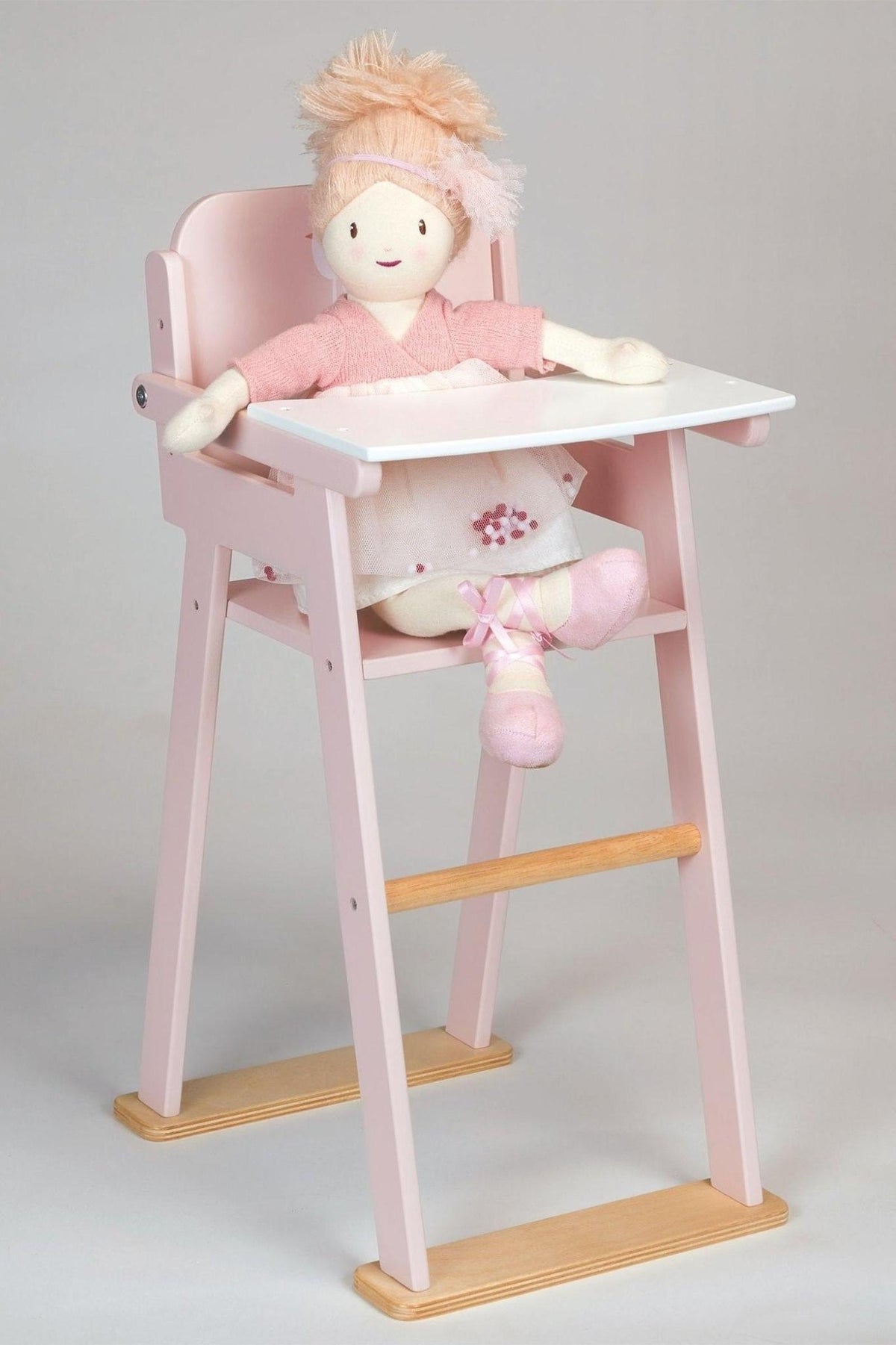 A fabric doll sits in a pink wooden high chair, wearing a pink outfit and ballet shoes.