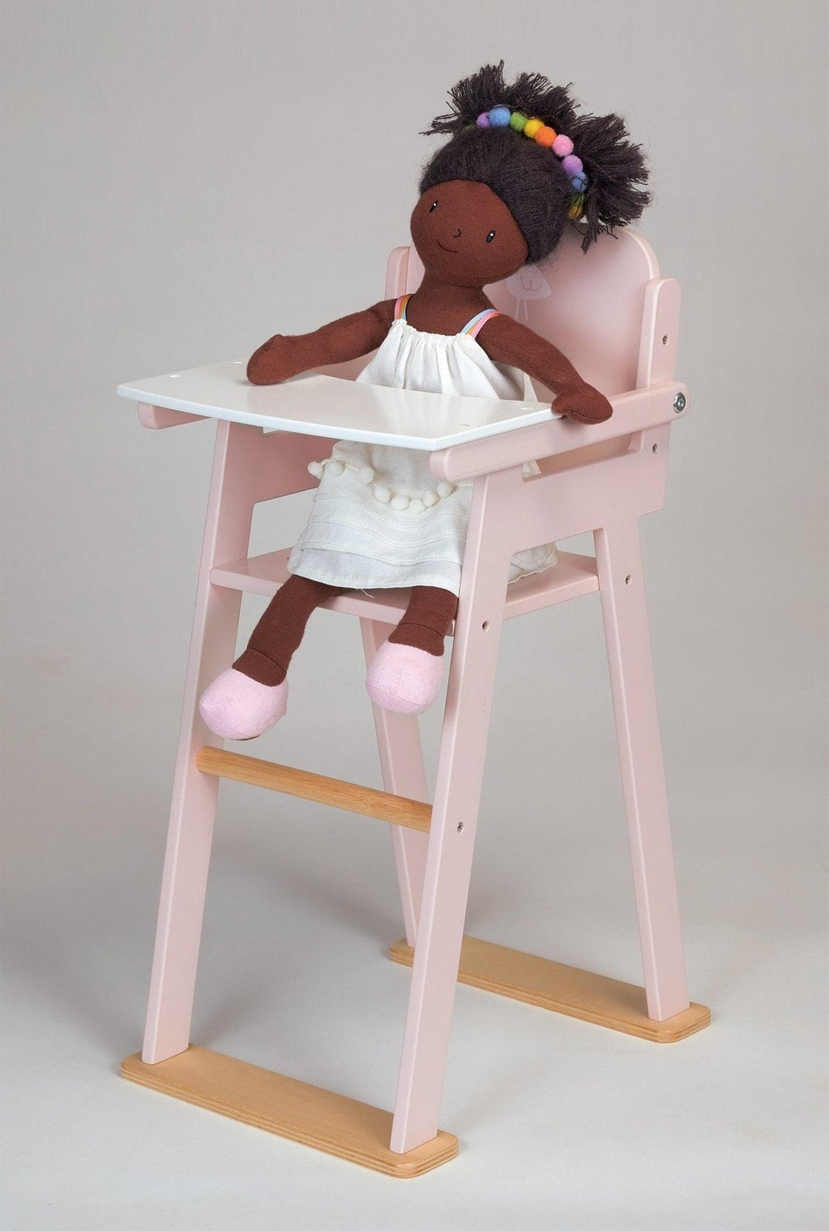 A doll in a white dress sits in a pink wooden high chair with a white tray.