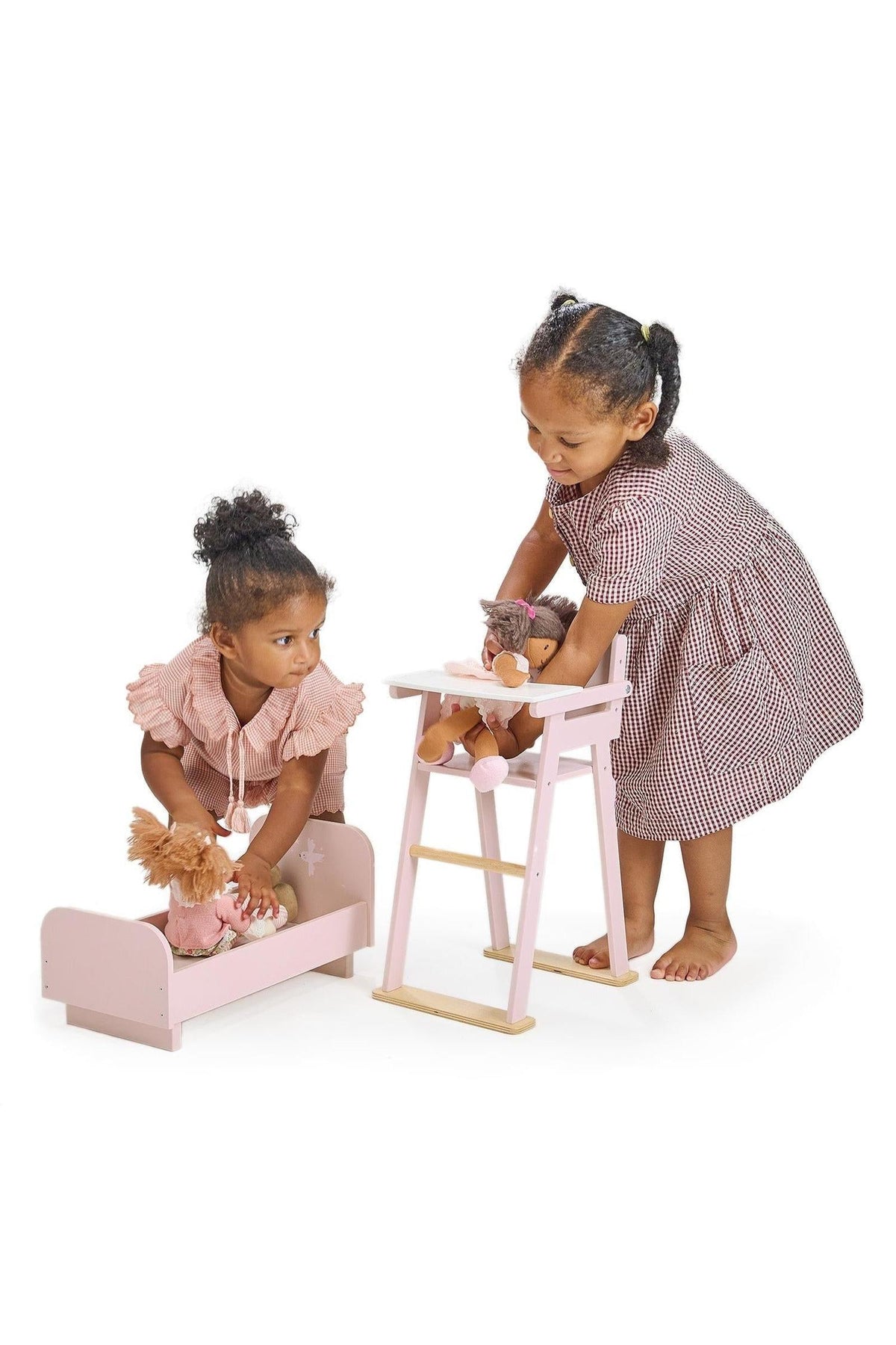 Two children play with pink wooden doll furniture, including a crib and high chair, on a white background.