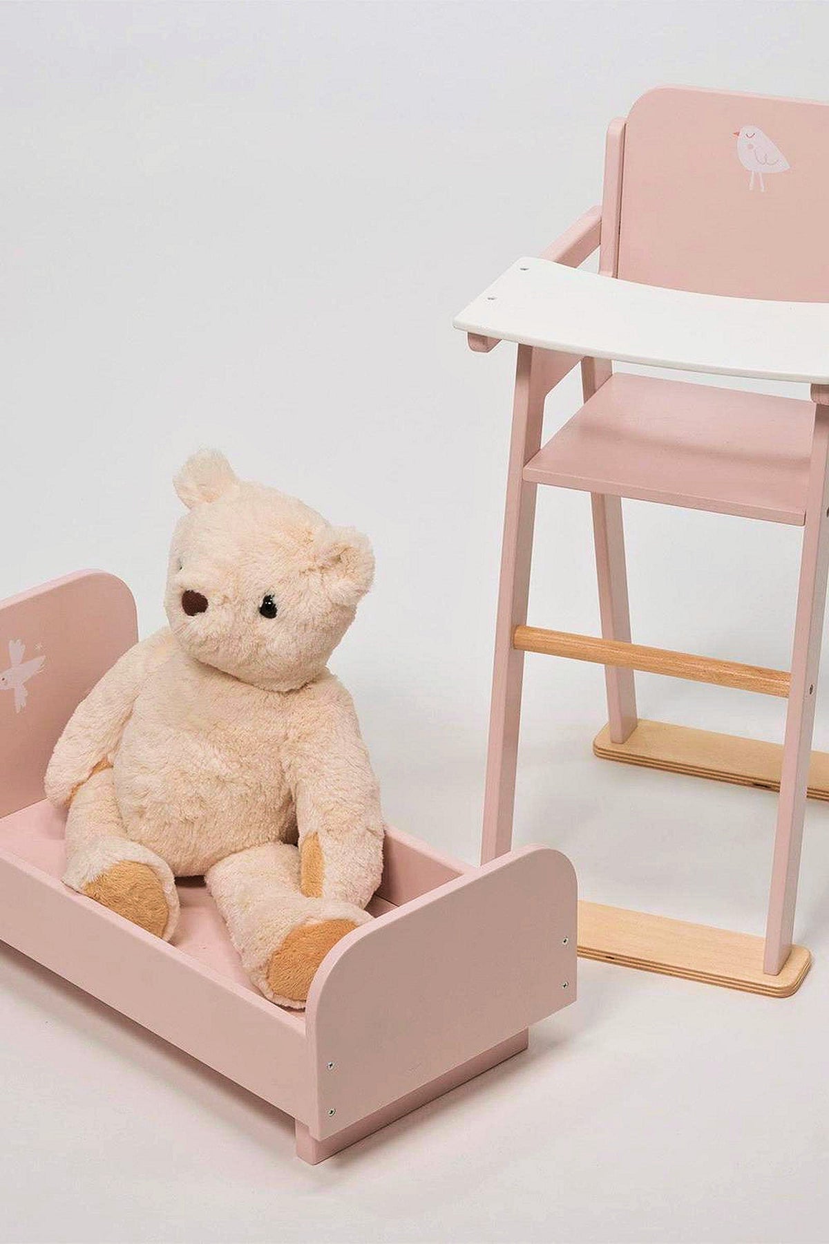 A plush teddy bear sits in a pink wooden crib next to a matching high chair on a white background.