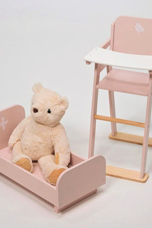 A plush teddy bear sits in a pink wooden crib next to a matching high chair on a white background.