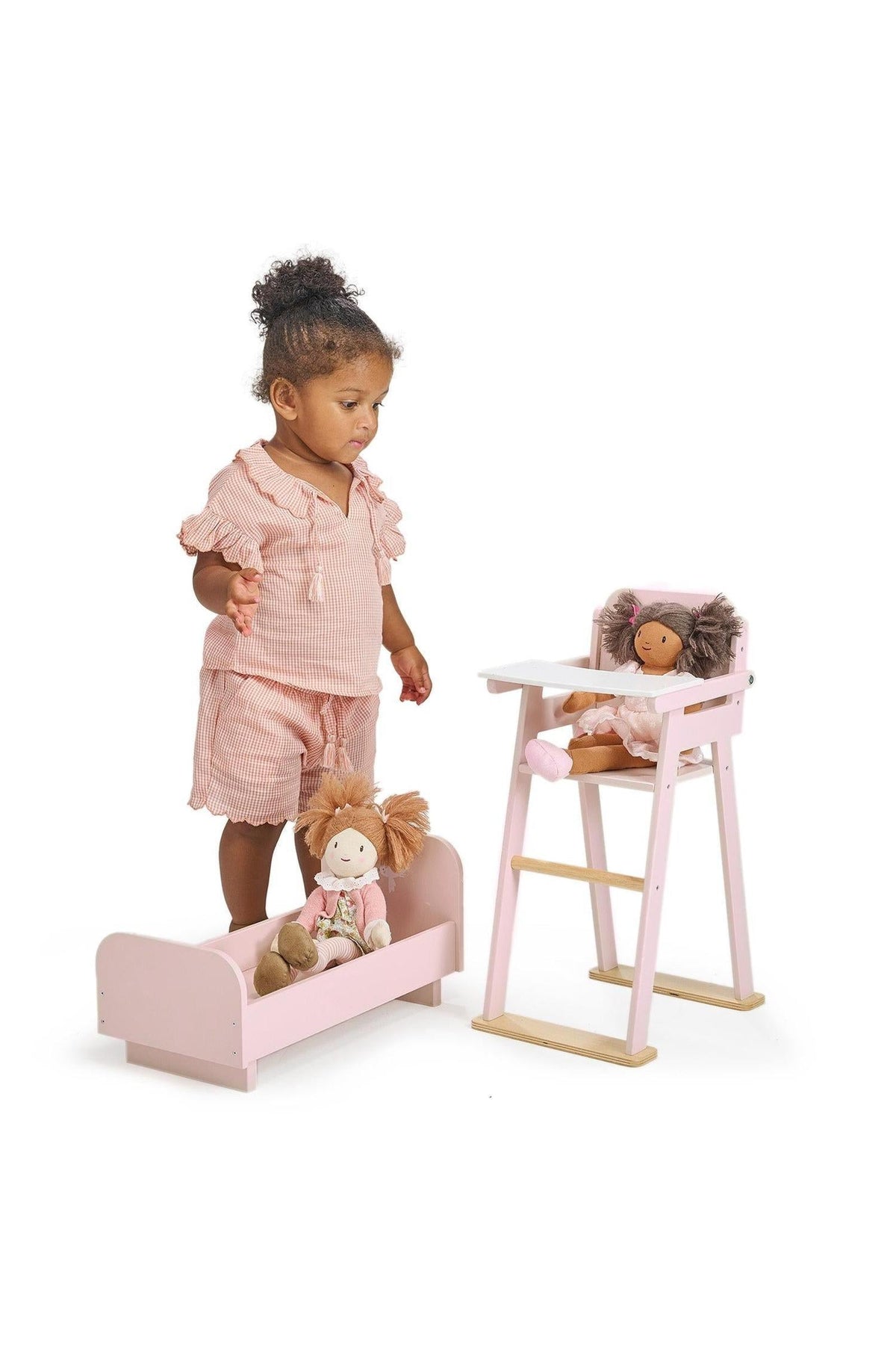 A toddler plays with a pink wooden doll crib and high chair set, featuring two dolls.