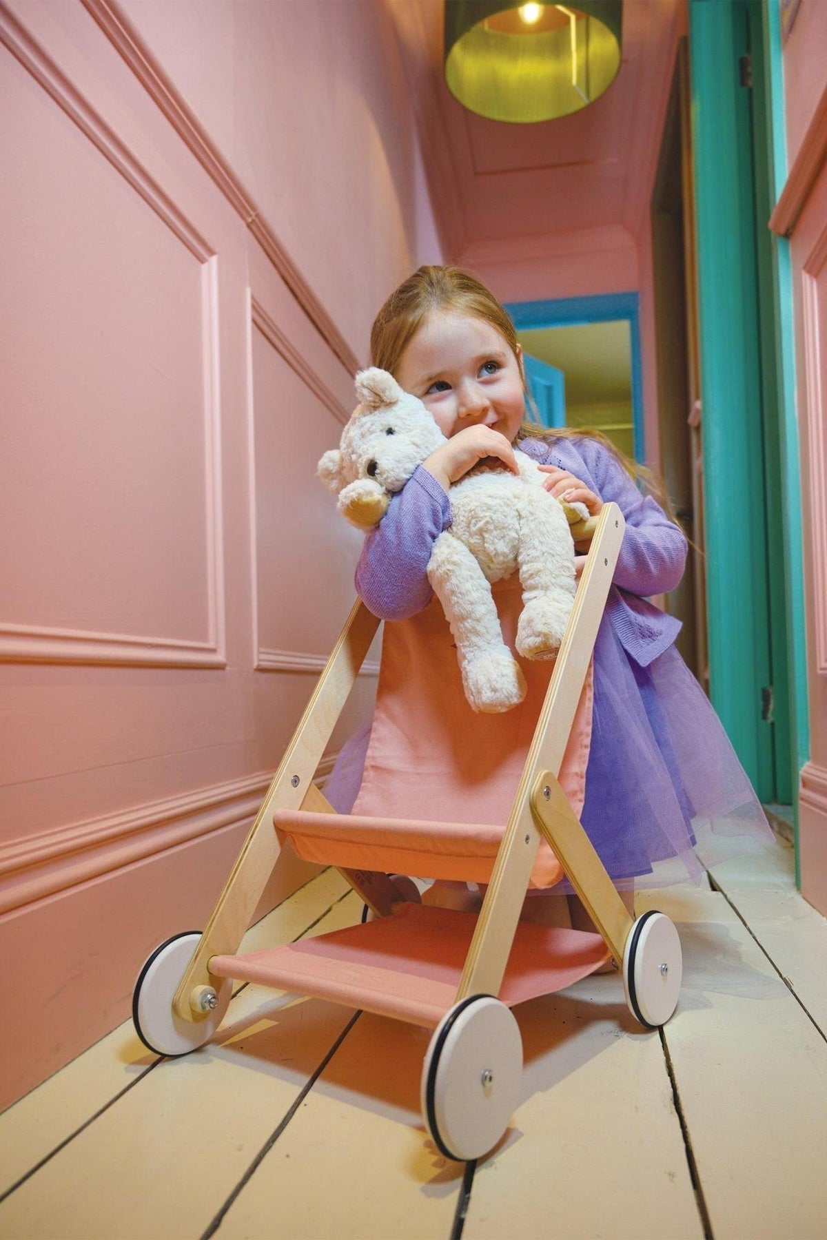 A child in a purple dress pushes a pink wooden stroller holding a stuffed animal in a pastel hallway.