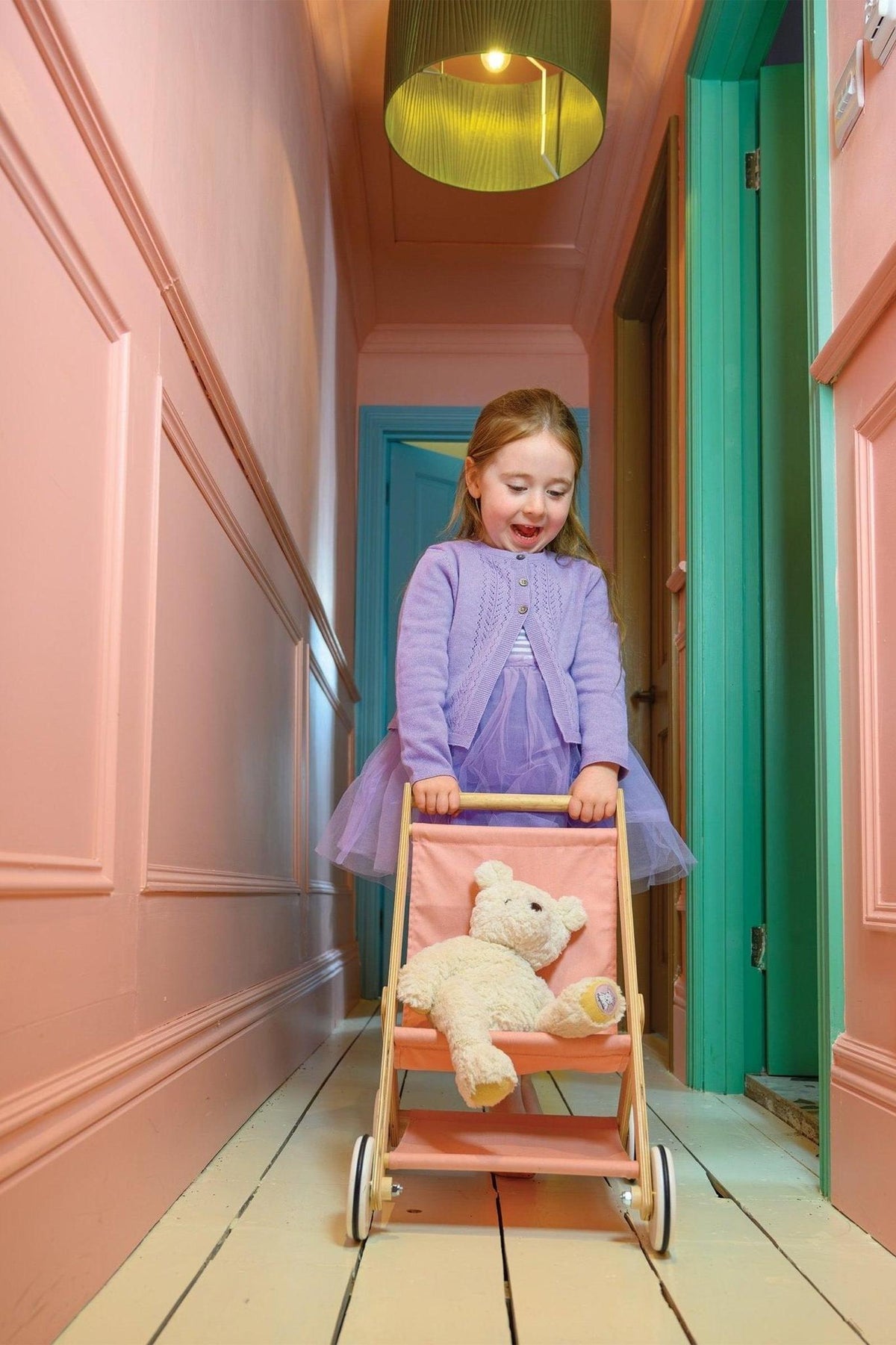 A child in a purple outfit pushes a wooden doll stroller with a teddy bear, in a colorful hallway.