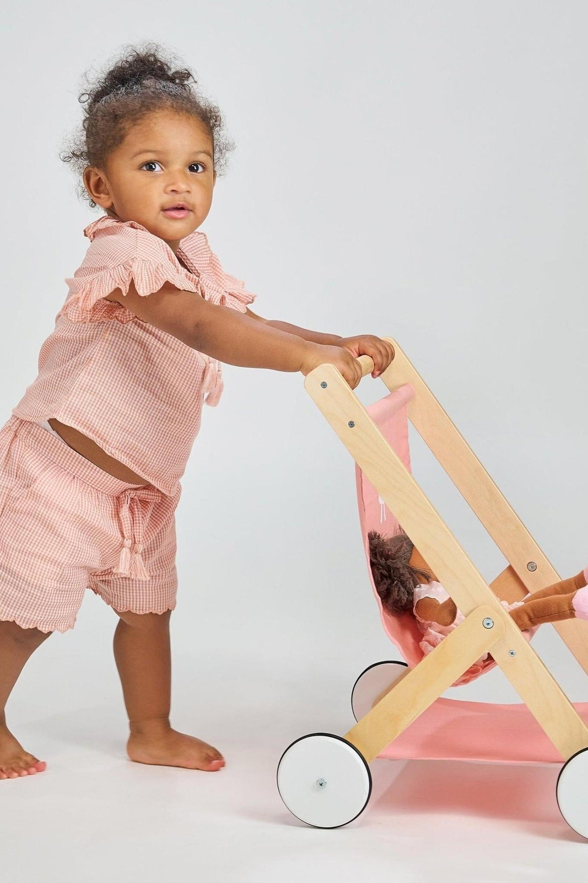 A toddler pushes a wooden baby doll stroller with a pink fabric seat, dressed in a pink outfit.
