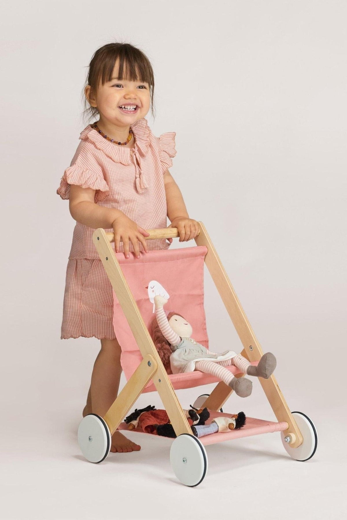 A smiling child pushes a pink and wooden doll stroller with a doll inside, wearing a pink dress.