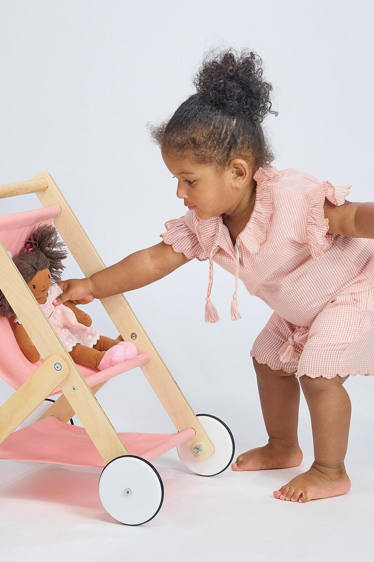 A toddler in a pink outfit plays with a wooden doll stroller holding a plush doll.