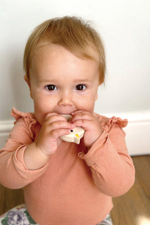 Baby in a pink shirt holding a white object, possibly a teething toy, against a plain background.
