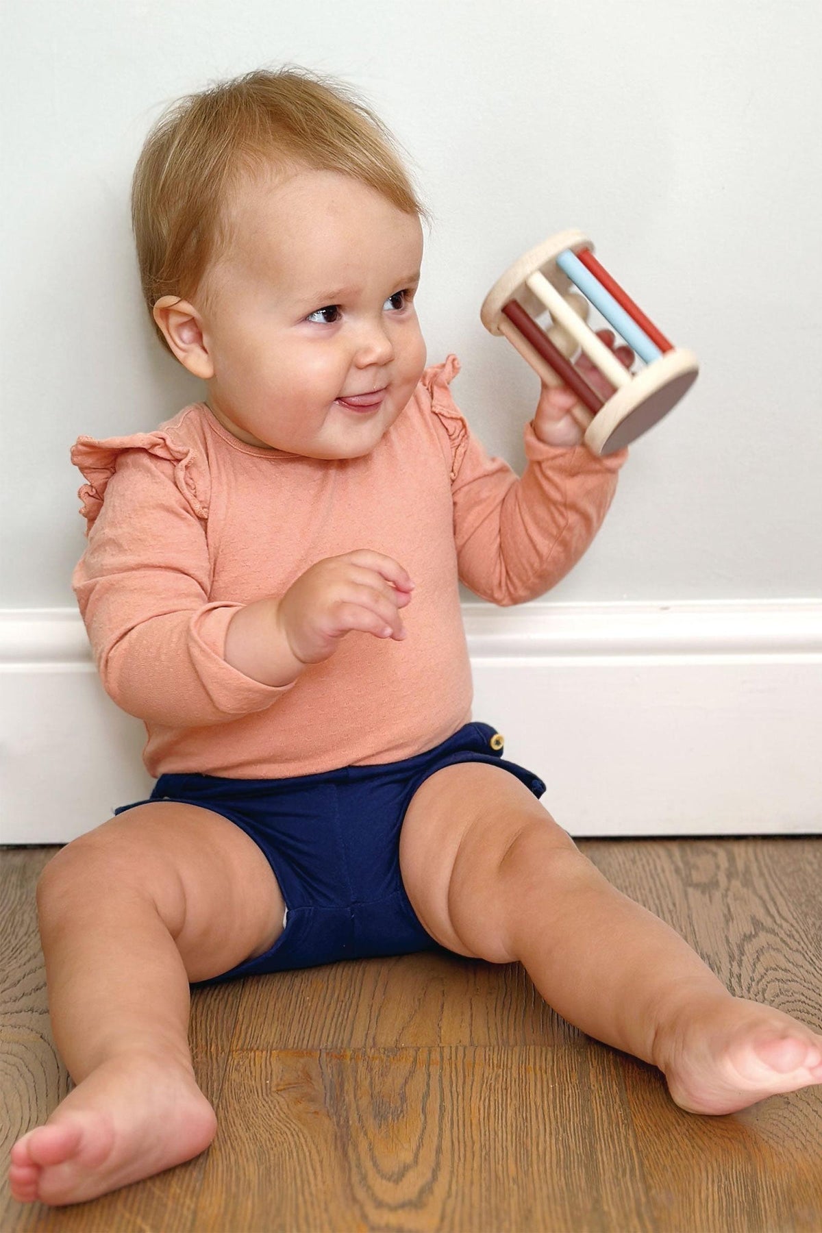Baby sitting on a wooden floor holding a toy, wearing a pink shirt and blue diaper.