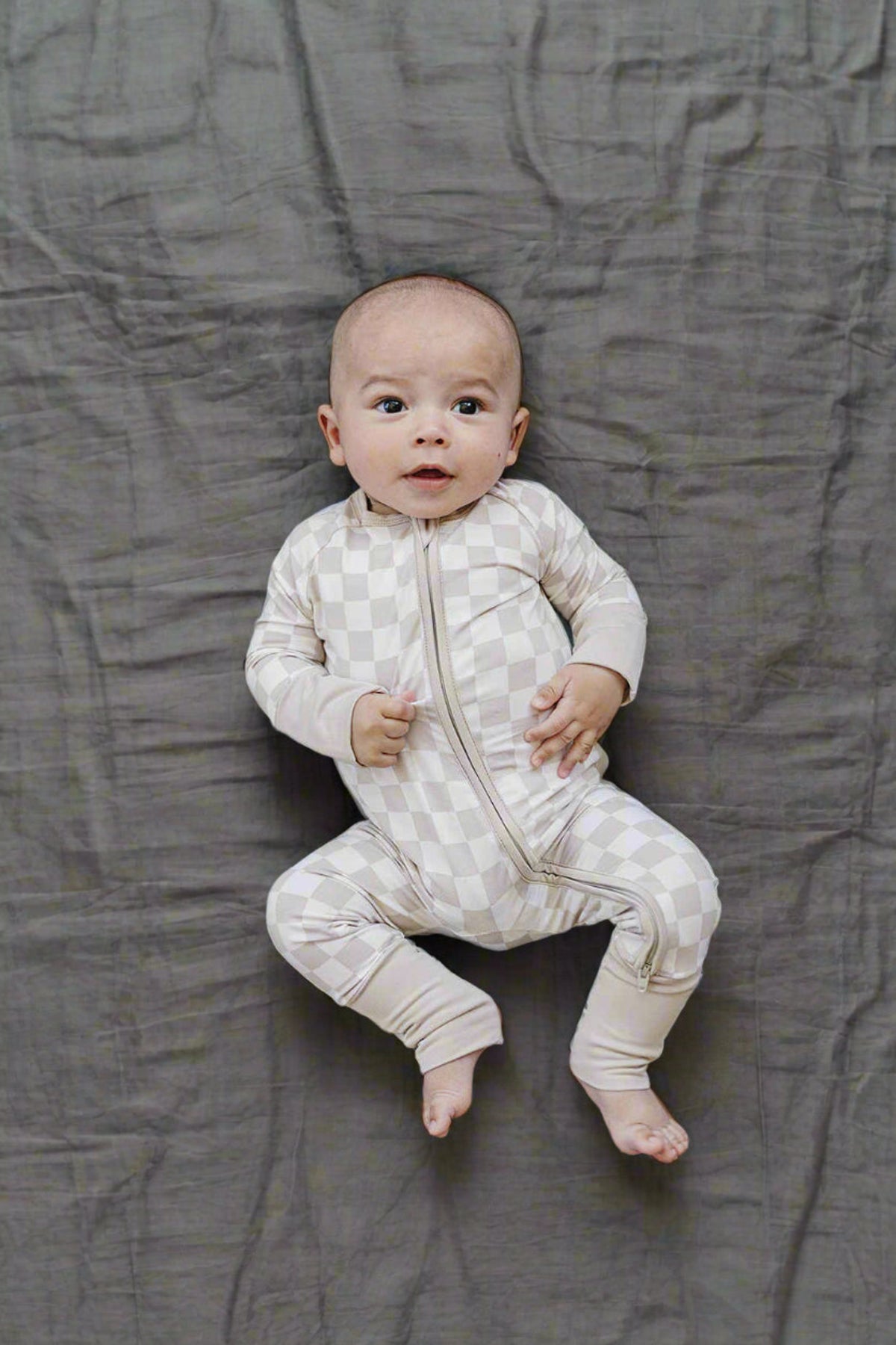 Baby in taupe checkered zipper pajamas made of bamboo fabric on a gray sheet. Pajamas have a double zipper.