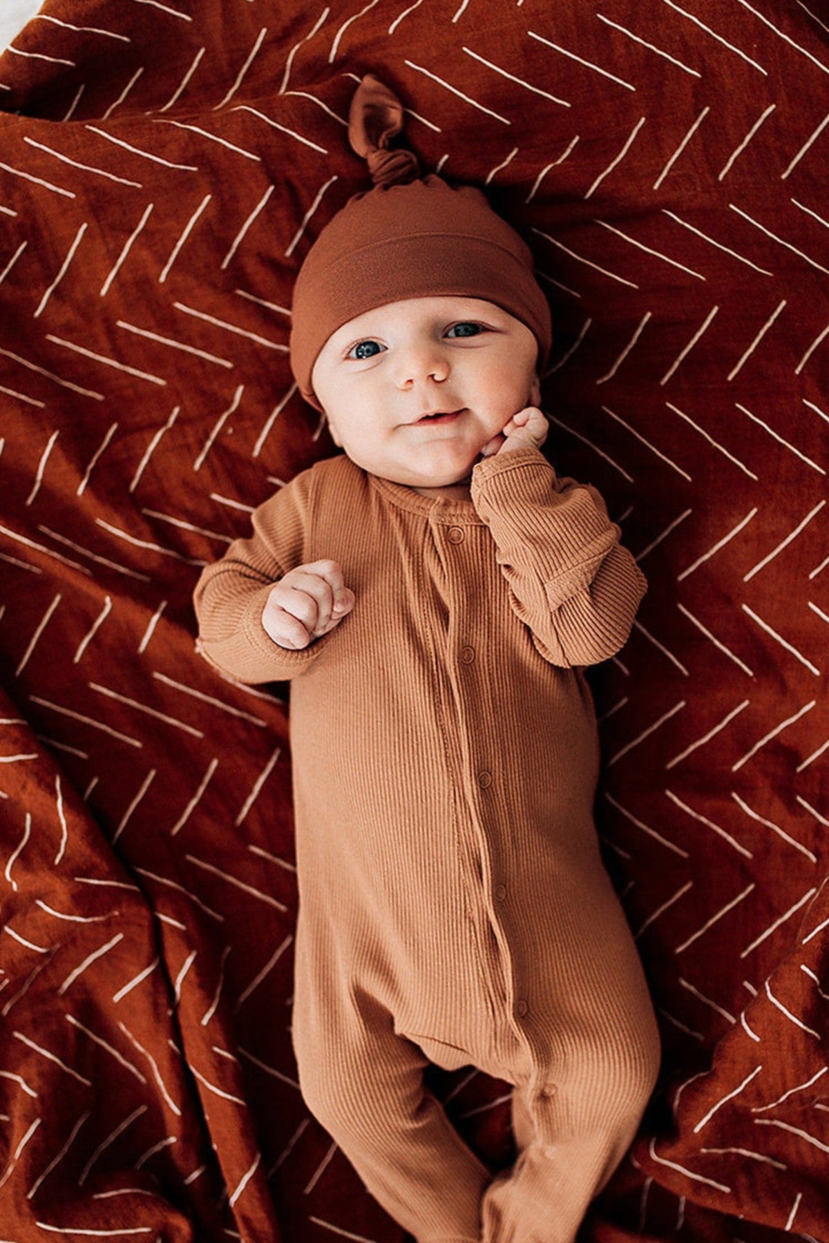 A baby wearing a rust-colored knot hat and matching onesie lies on a patterned blanket.