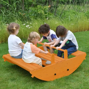 Four children play together on a vibrant orange wooden rocker in a lush green outdoor setting, enjoying active playtime on this sturdy, eco-friendly climbing toy designed for toddlers.