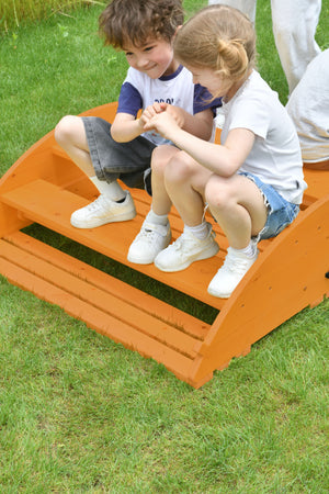Two children sit together on a bright orange wooden Jumbo Rocker and Climber, enjoying playful interaction on a grassy outdoor setting, showcasing its sustainable design for active play.