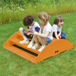 Three children play on a bright orange wooden Jumbo Rocker and Climber set, sitting and laughing on the grassy outdoor surface, enjoying active, imaginative play in a natural setting.
