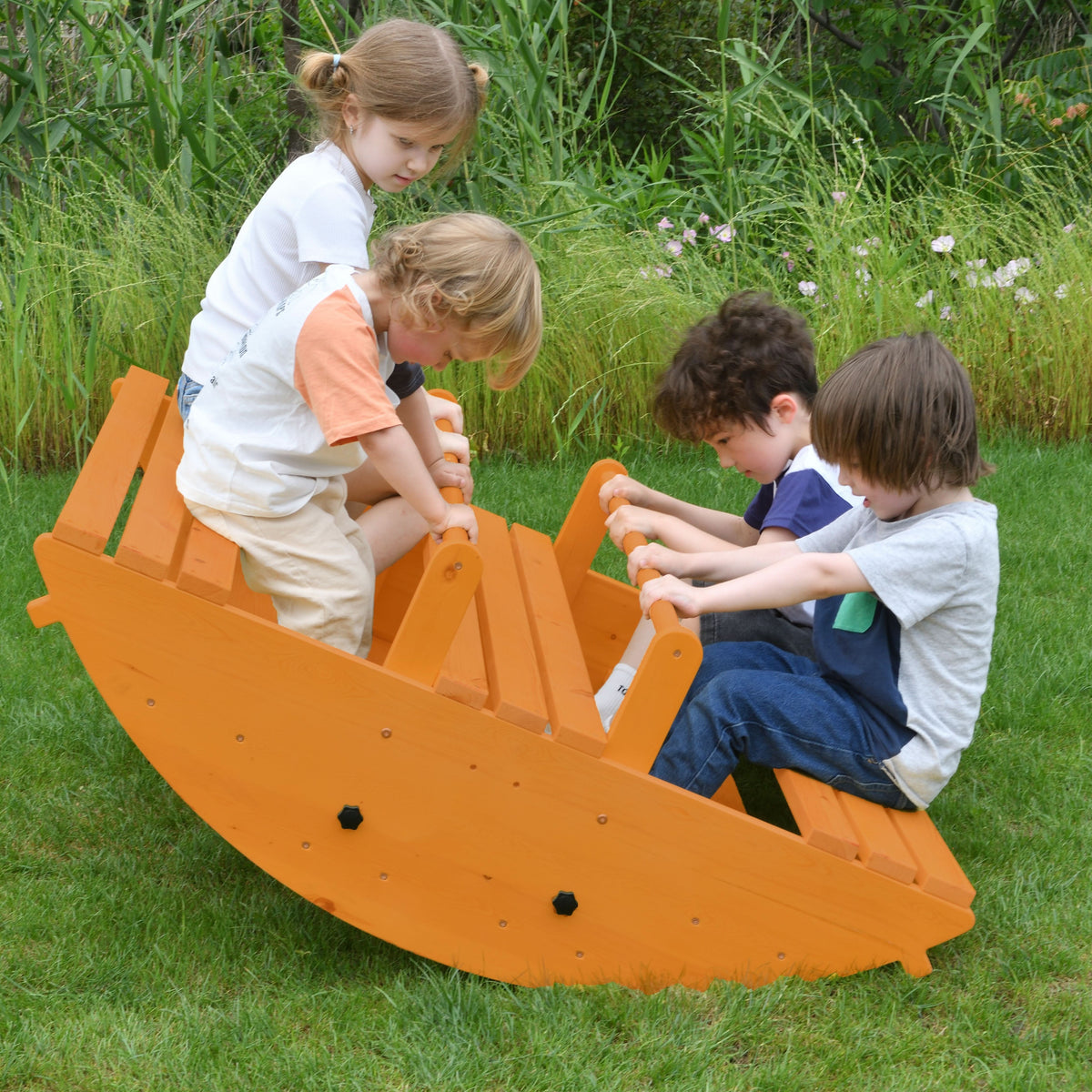 Four children play together on a bright orange wooden rocker, engaging in active play on a grassy outdoor setting. The oversized design encourages climbing and balancing for toddlers.