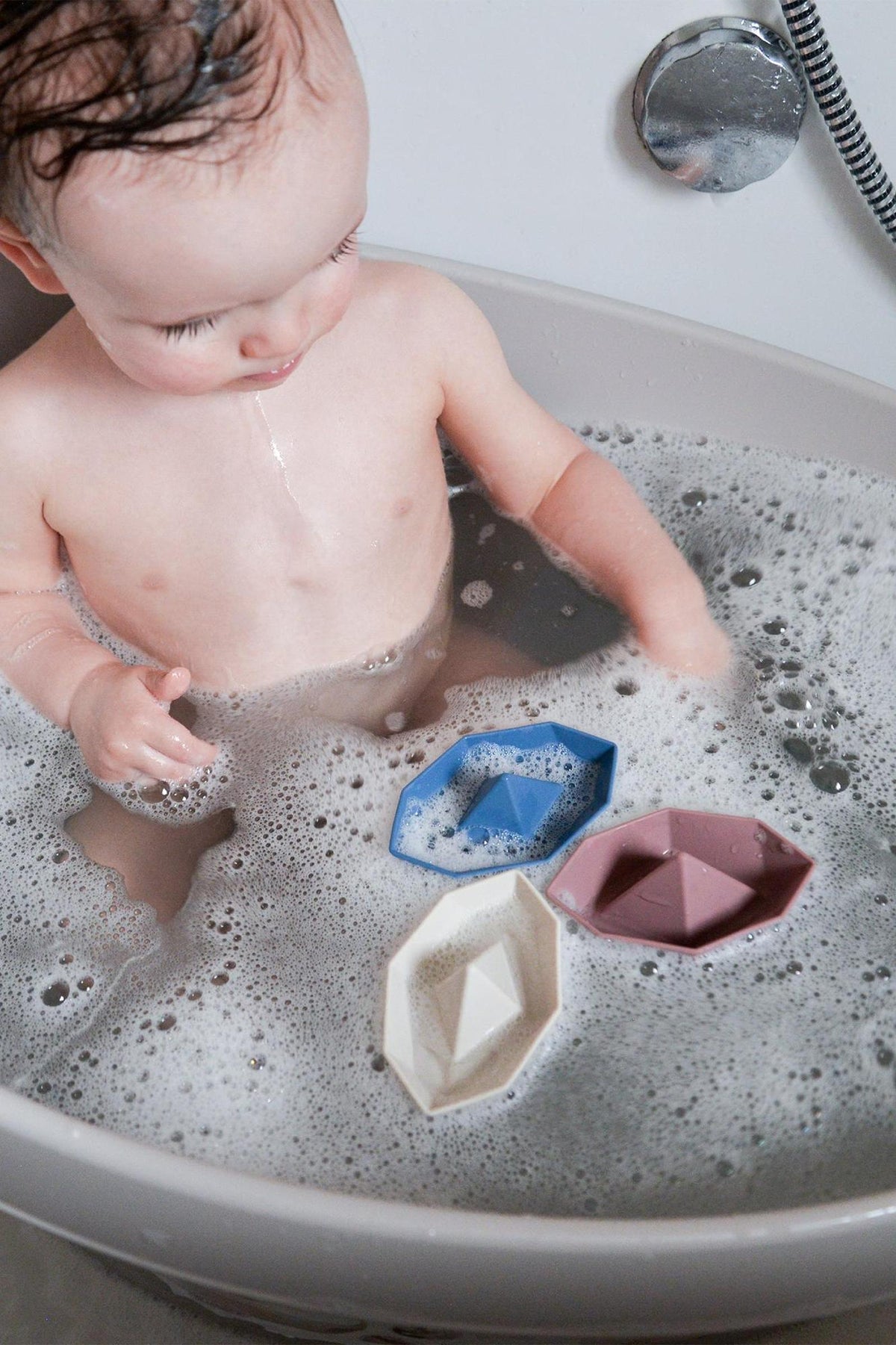 A baby in a taupe bathtub plays with colorful toy boats in soapy water, featuring blue, pink, and cream.